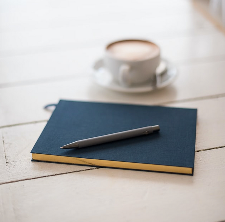 A closed black journal with a pen laying on cover. The journal is sitting on wood table next to a small cup of coffee.   